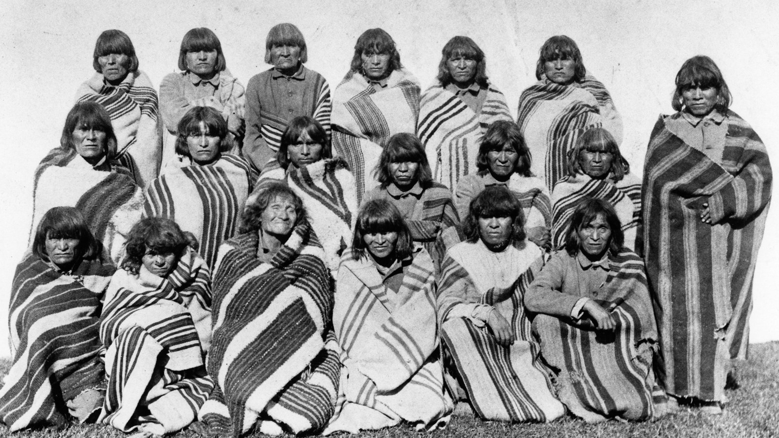 Black and white image of Hopi Indians arranged in two rows. They are wrapped in a striped blanket.