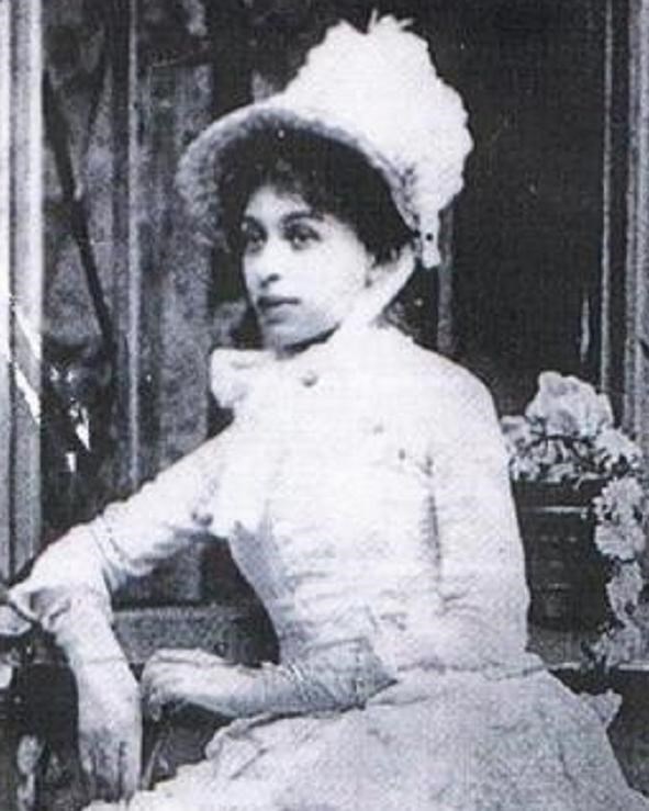 A young Black woman sitting for a portrait wearing a long-sleeved white dress and large hat.