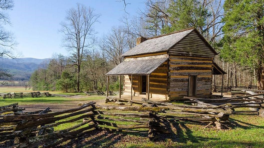 Historic cabin located in an open meadow behind a split rail fence with trees behind it.s