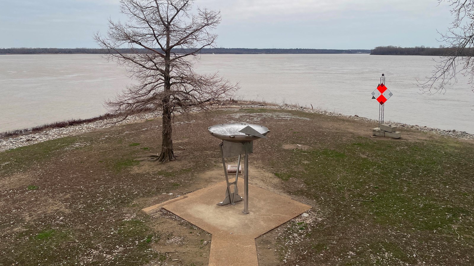 a paved walkway leads to a mounted metal viewing scope aimed toward the wide, muddy river.