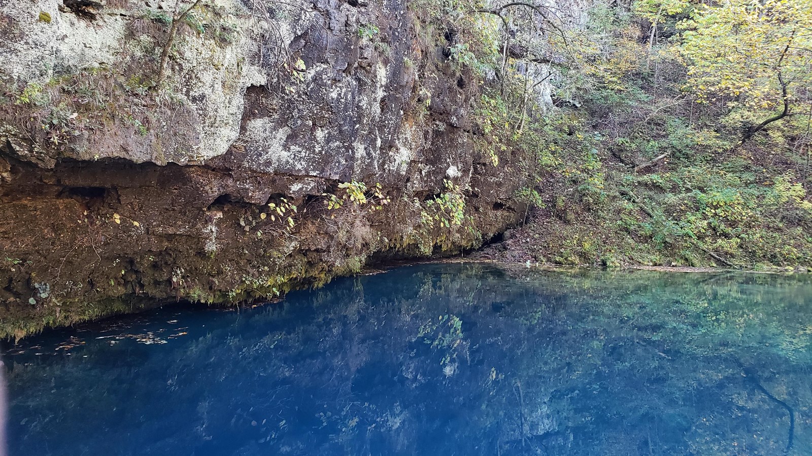 A dark blue pool of water with a grey rock bluff behind it with green vegetation on rocks