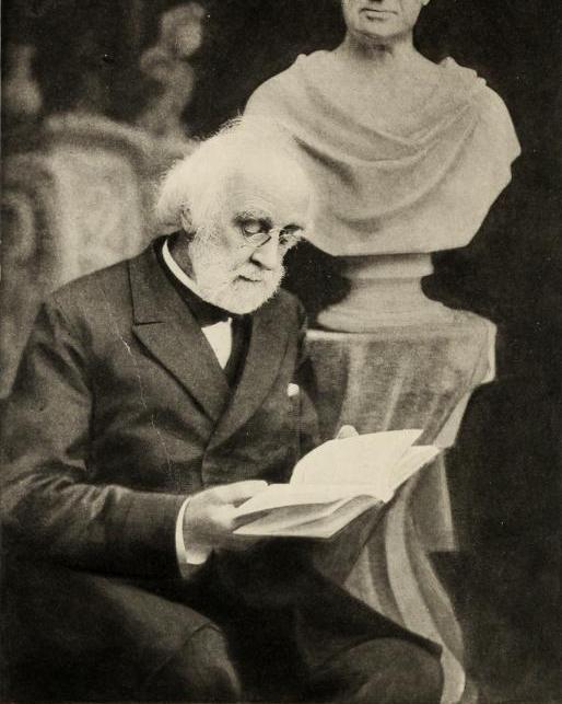 Portrait of an older man sitting reading an open book in front of a bust 