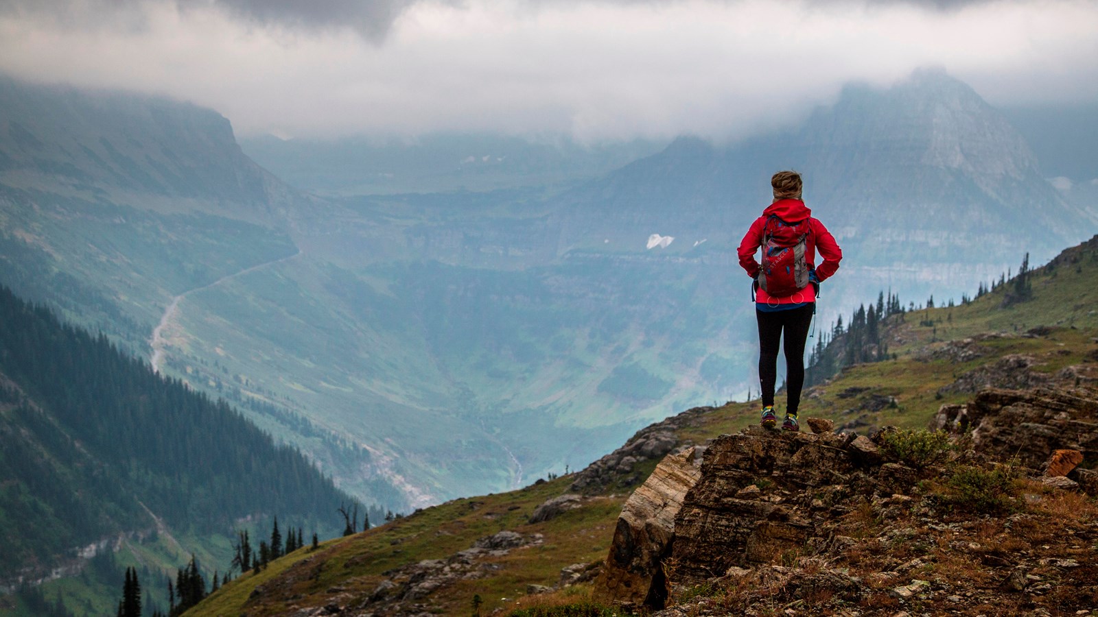 A hiker in red takes in a sweeping landscape cloaked in grey clouds.