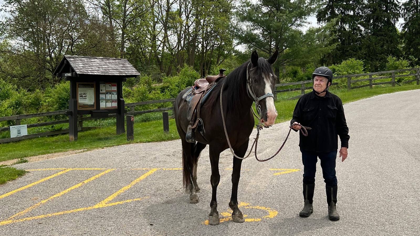 A man in a helmet and his brown horse stand in the lot near a trailhead bulletin board.