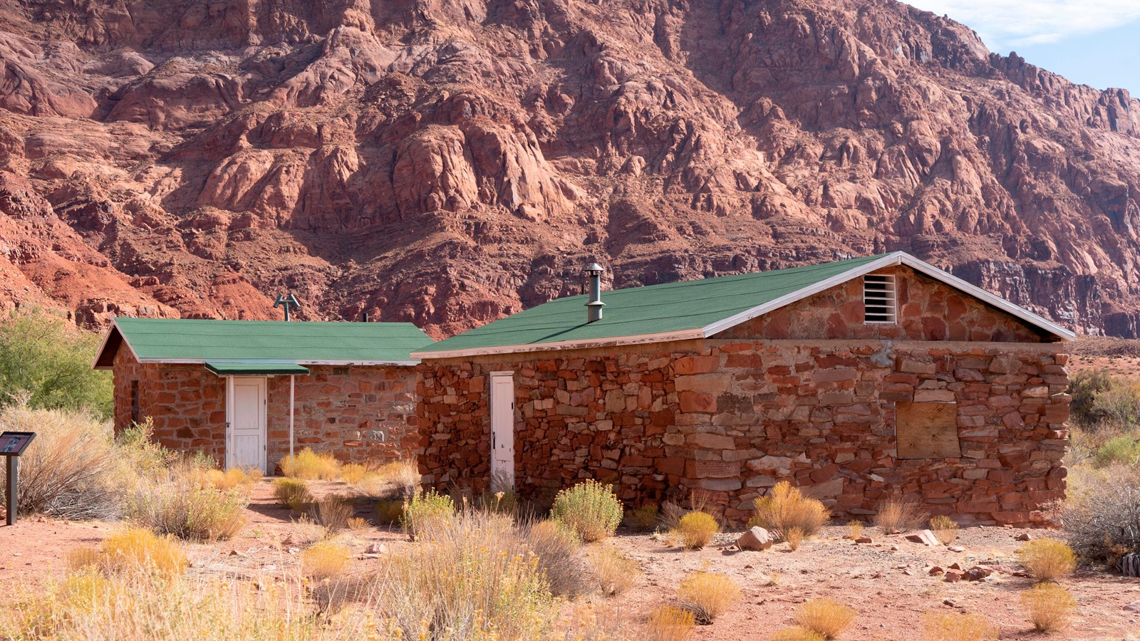 Interpretive sign in front of two stone buildings with white wooden doors and a modern green roof.