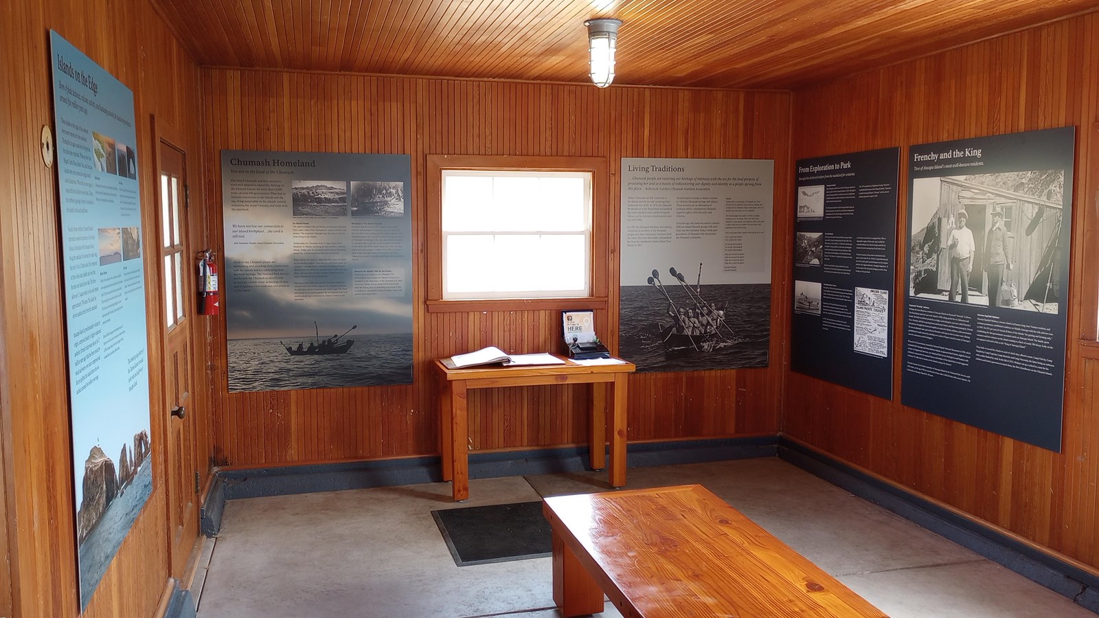 wooden walled interior building with cement floor, exhibit signs on the walls, and a bench and table