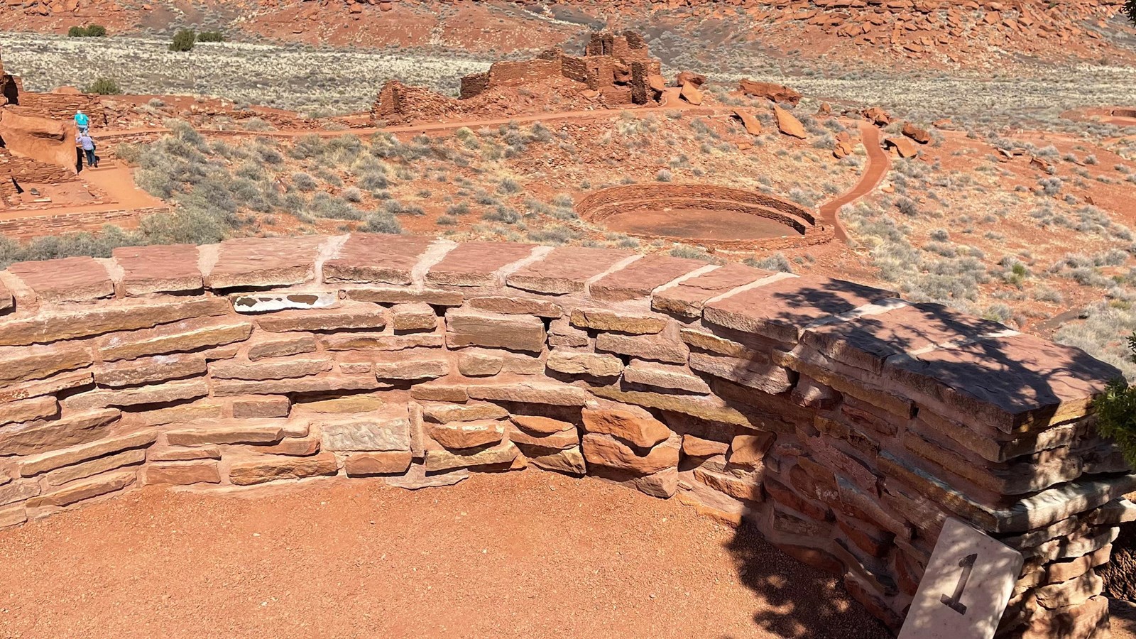 A trail overlook bordered with a low sandstone wall and a large stone pueblo in the background.