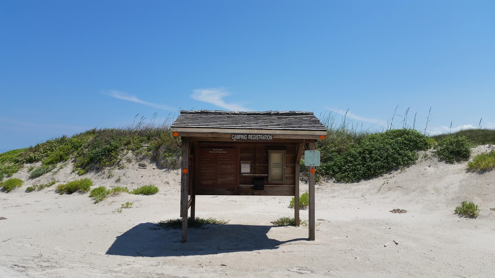 A wooden information kiosk on the beach with sand dunes behind.