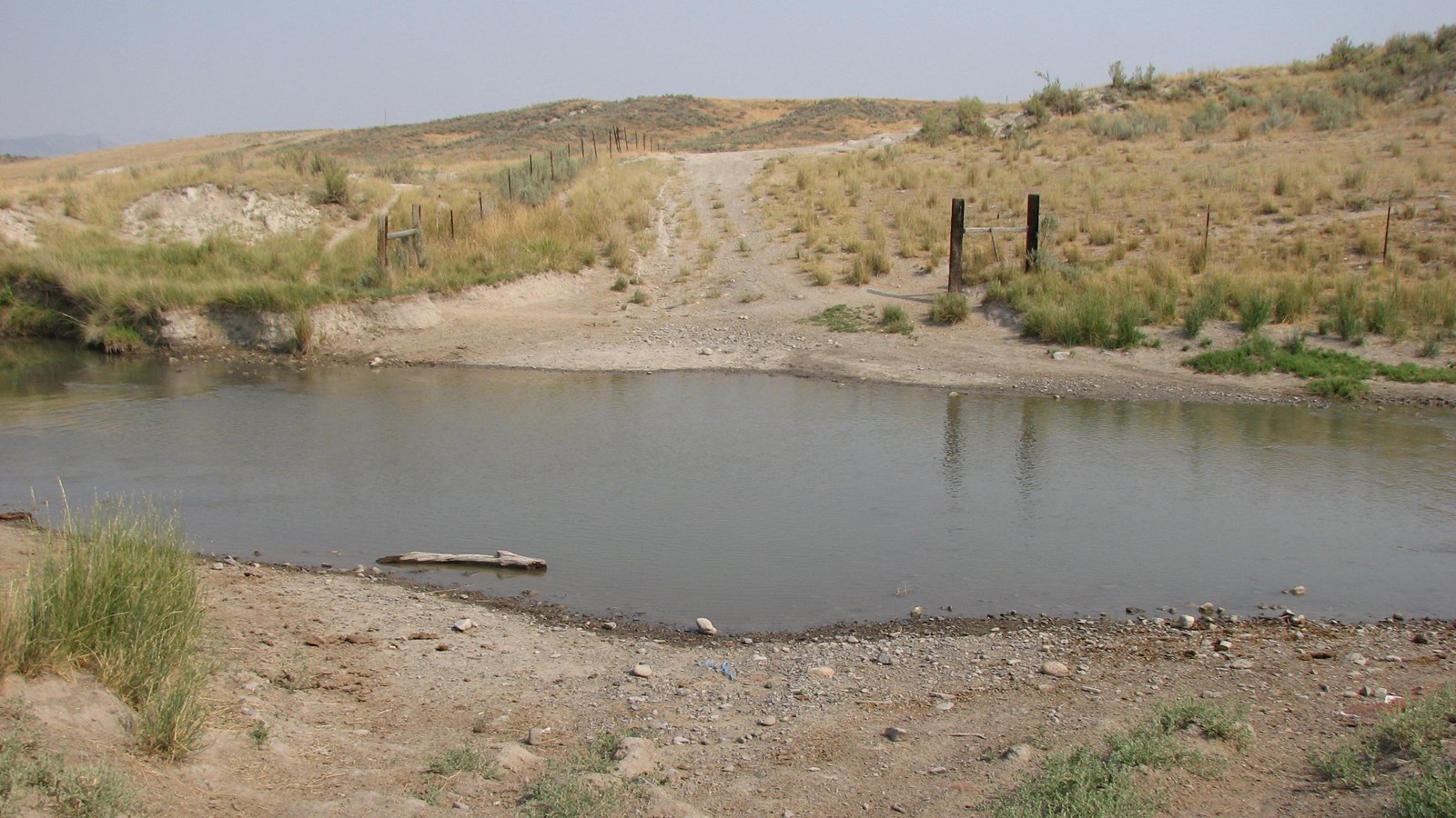 In the foreground, an apron of dirt and gravel leads to the murky green waters of the Malad River.