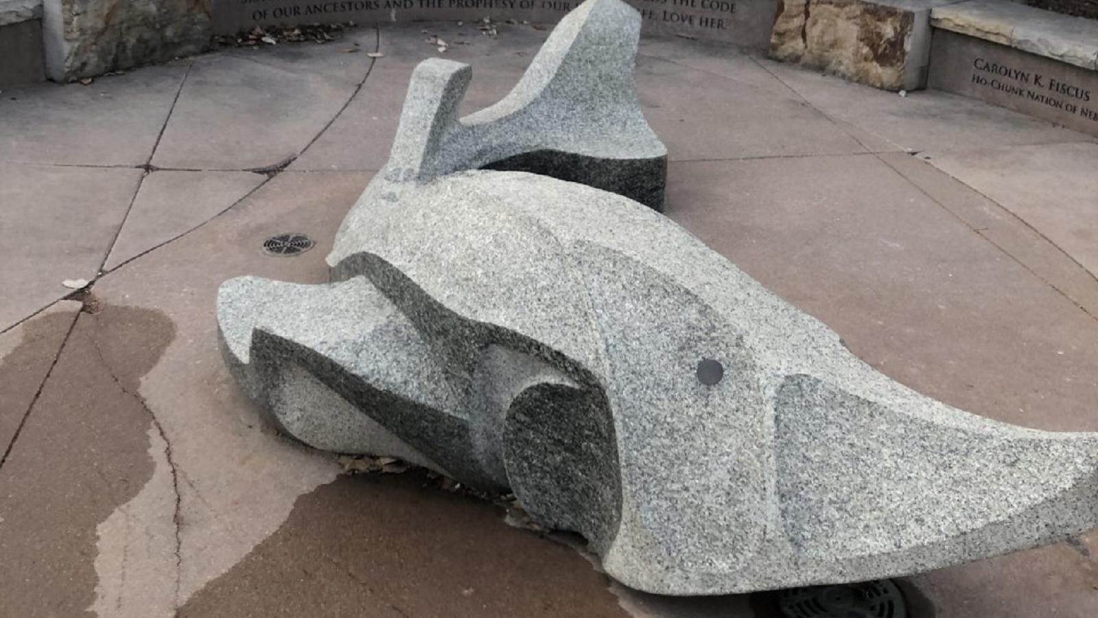 A statue of a sturgeon made of speckled tan granite lays atop a paved patio space