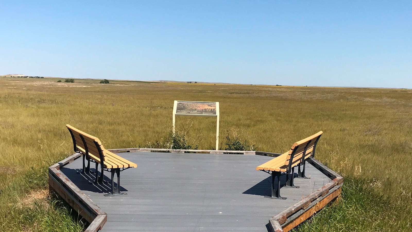 Prairie grasses extend into the horizon under a blue sky, a boardwalk with two benches a wayside.