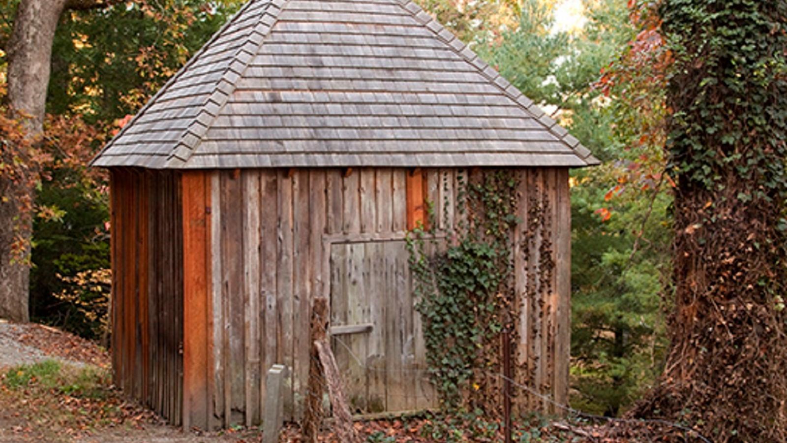 A square wooden building with wood shingles and a door sits in a wooded area