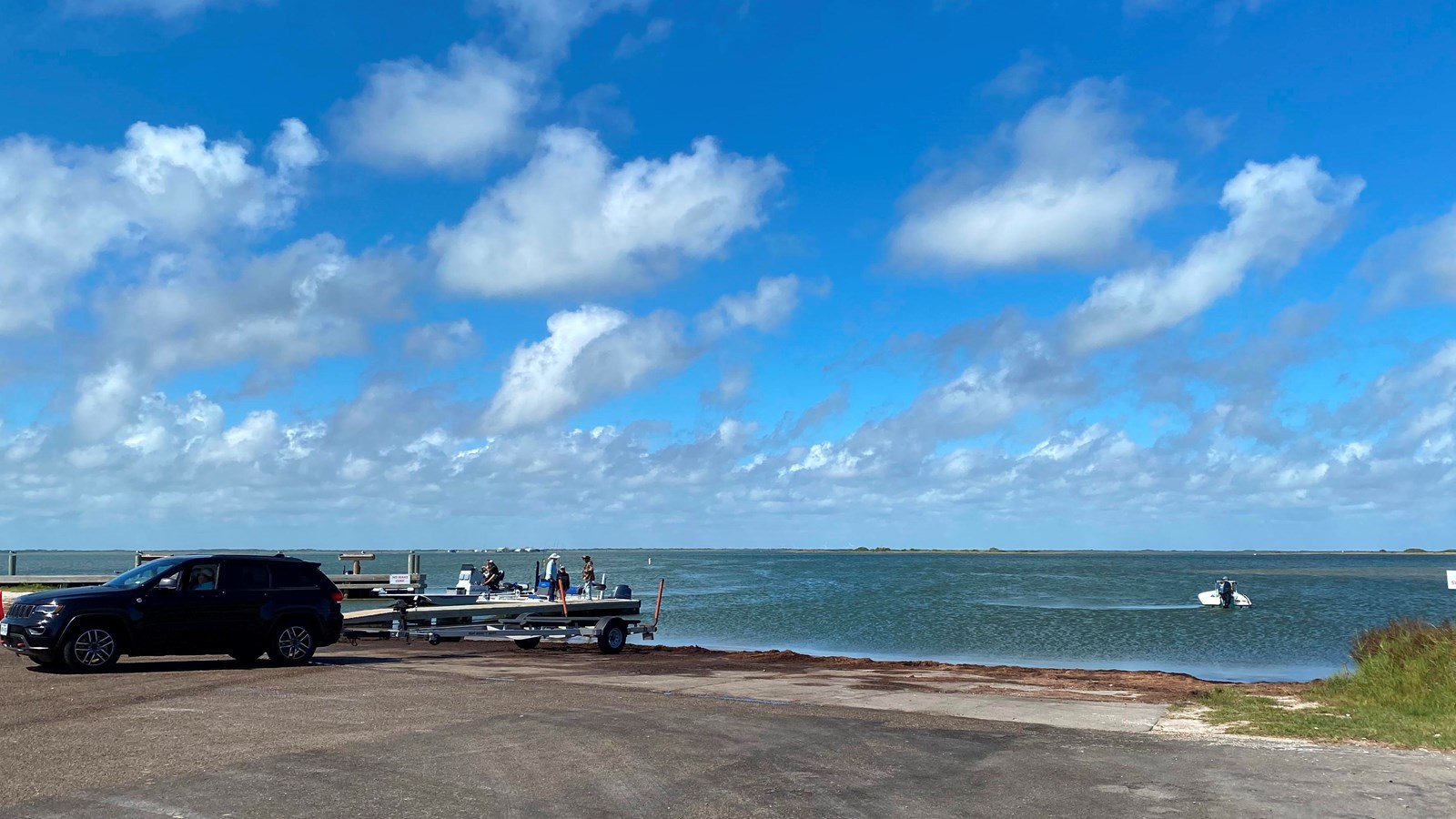 A black car towing a trailer backs down a ramp to the water. 