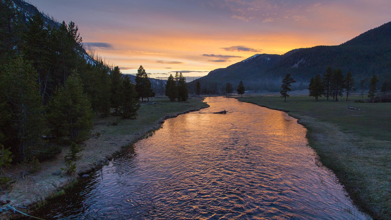 Purple, pink, and orange are cast across the sky and reflect in the slowly moving water of a river.