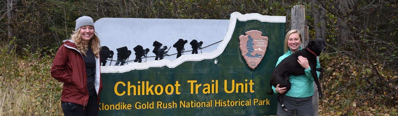 two women and a dog pose with a Chilkoot Trail sign