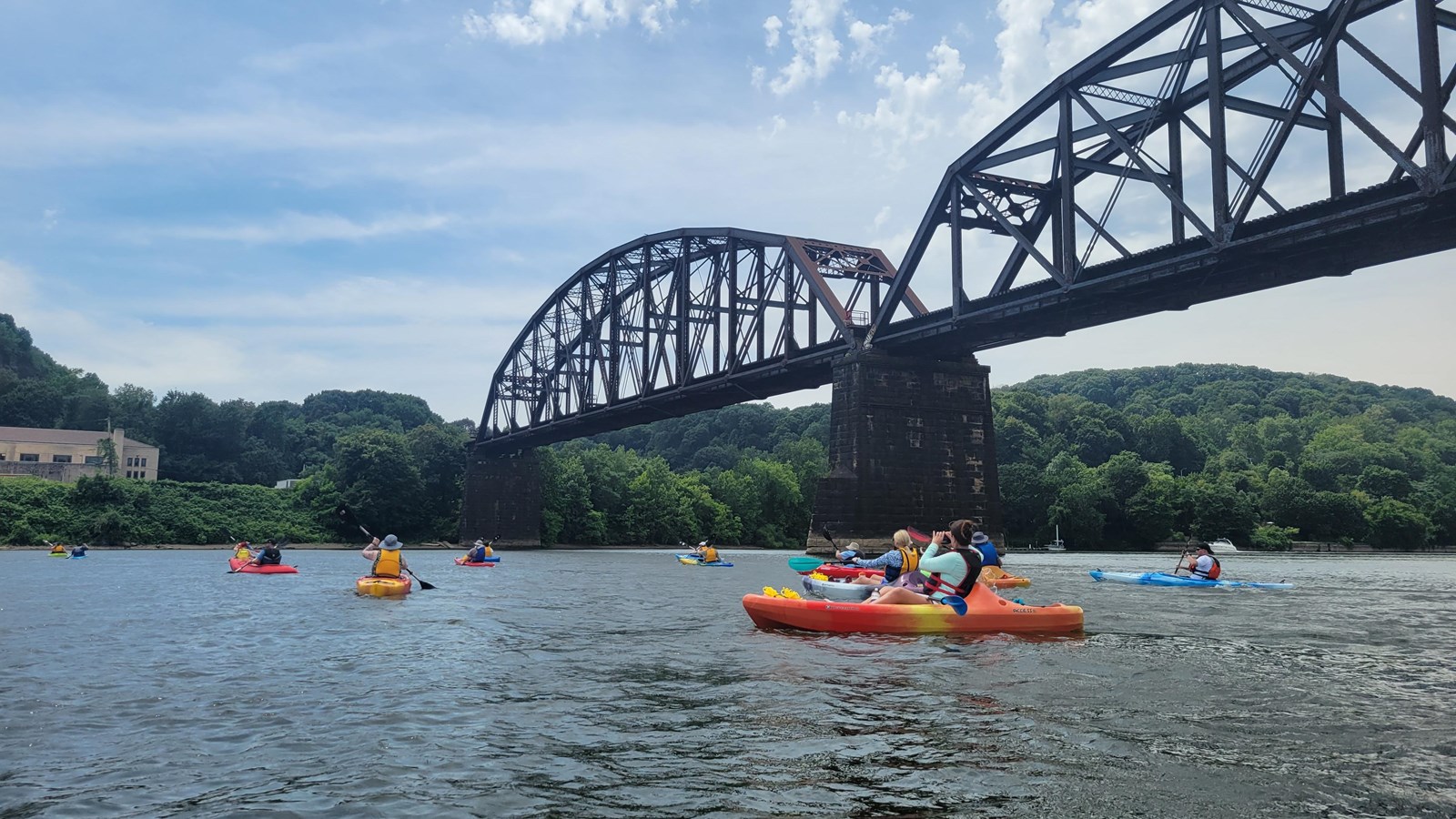 The photo shows a group of people kayaking on a broad river beneath a large steel truss bridge. 