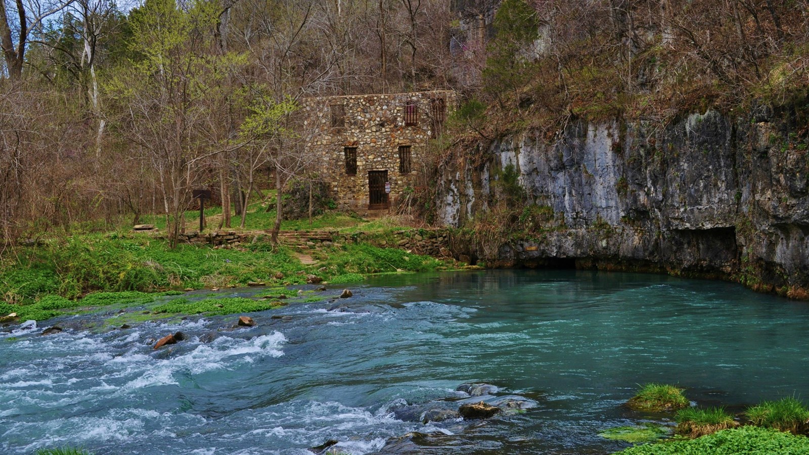 A blue pool of gushing water comes from a grey rock cliff to right, a stone structure is behind