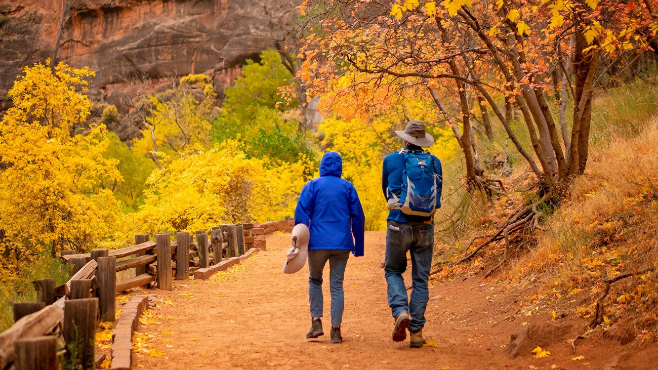 Two hikers in blue jackets walk along a sandy trail lined with a wooden rail and fall foliage.