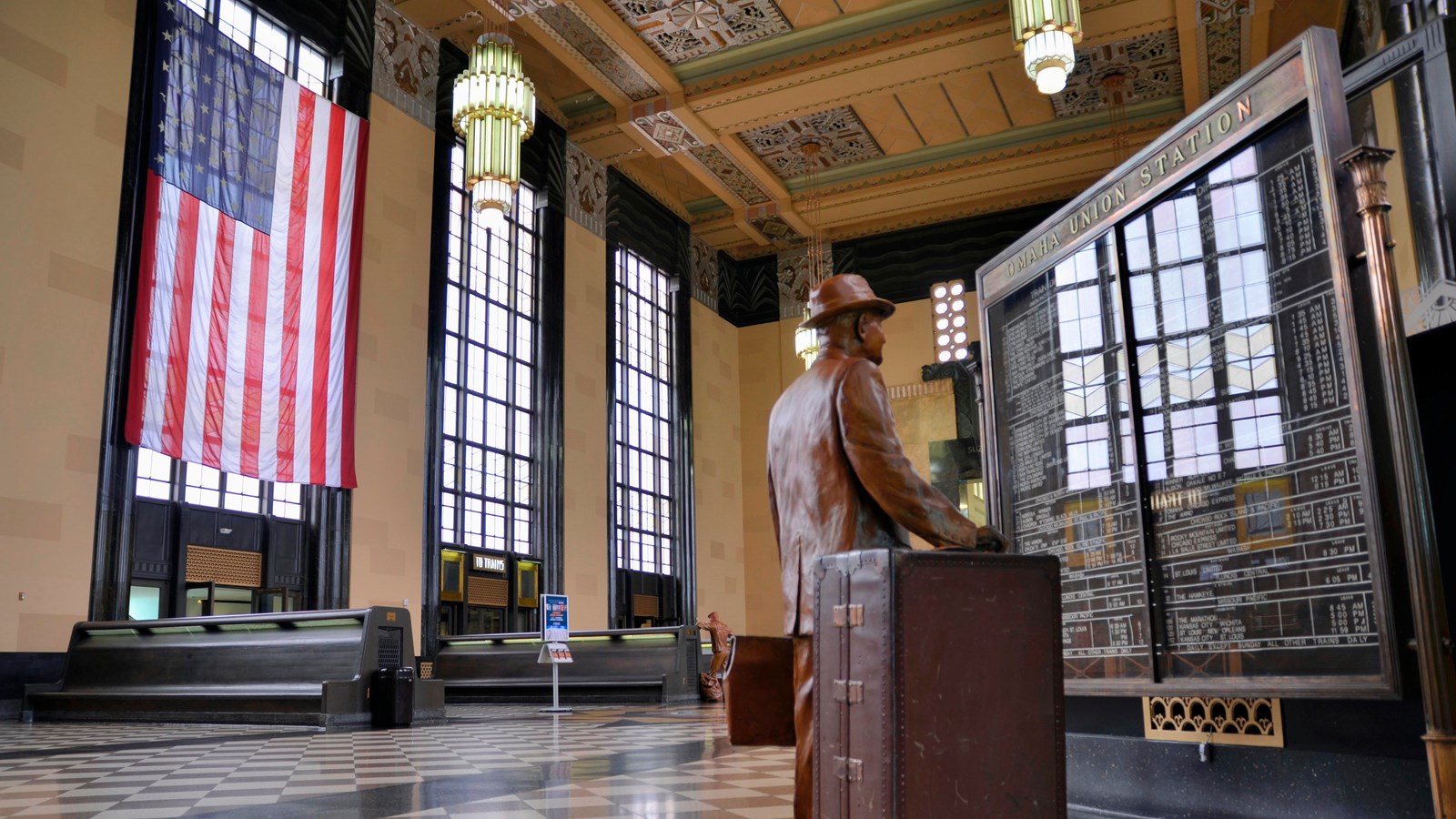 An ornate passenger train hall with large windows, a statue of a passenger and luggage