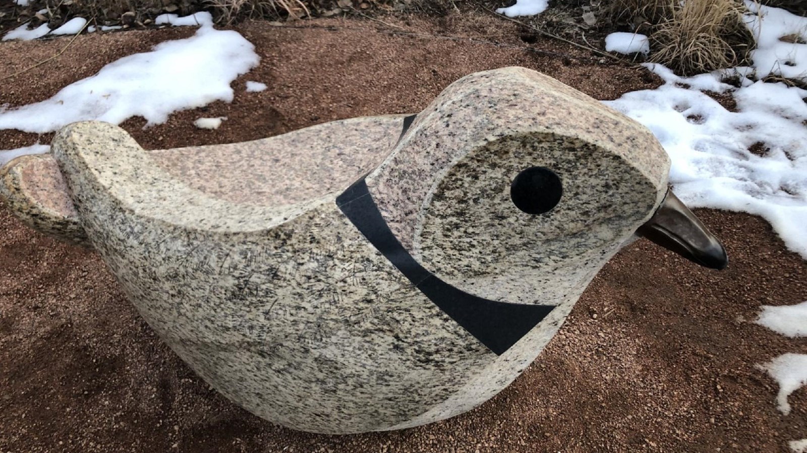 A statue of a bird carved from tan and black speckled granite sits atop pea gravel.