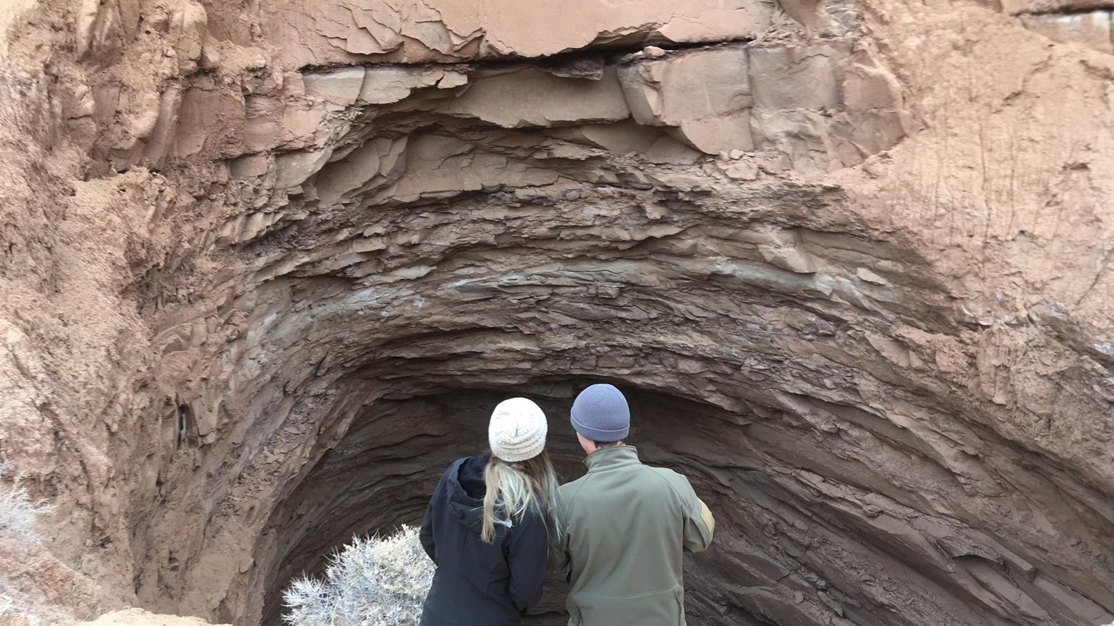 Two people stand at edge of large deep hole at base of reddish cliff.
