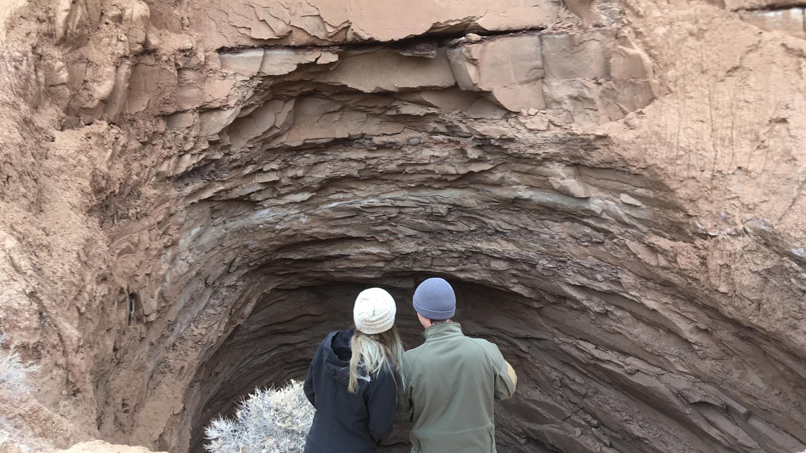 Two people stand at edge of large deep hole at base of reddish cliff. 