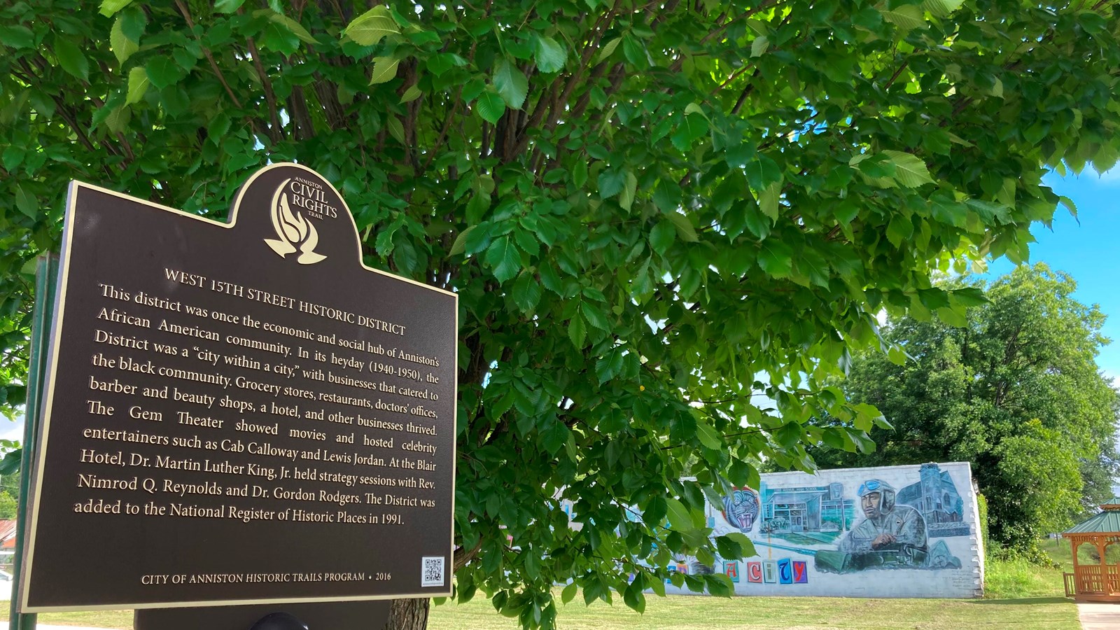 Foreground: a brown and gold historic marker. Background: trimmed green grass and a colorful mural.