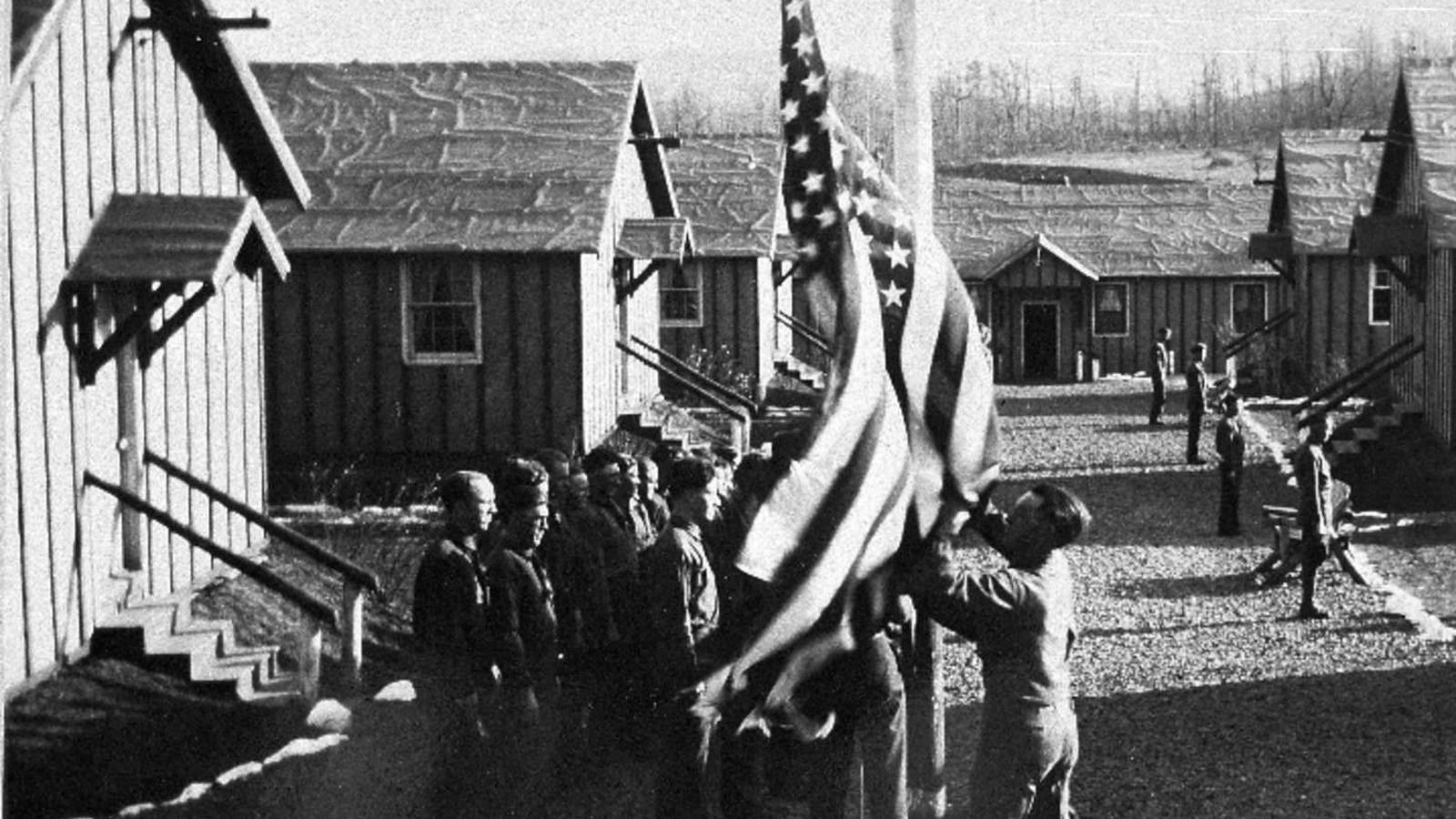 CCC boys wearing their dress uniforms stand at attention outside their barracks lowering a flag.