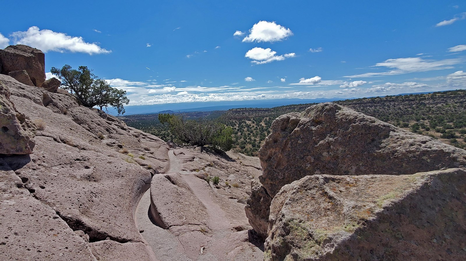 a carved trail crosses tan rock with distant views of mountains & trees, blue sky with white clouds