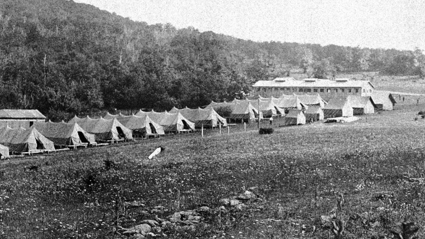 A wide angle view taken from a distance, of a CCC Camp. There are many large tents.