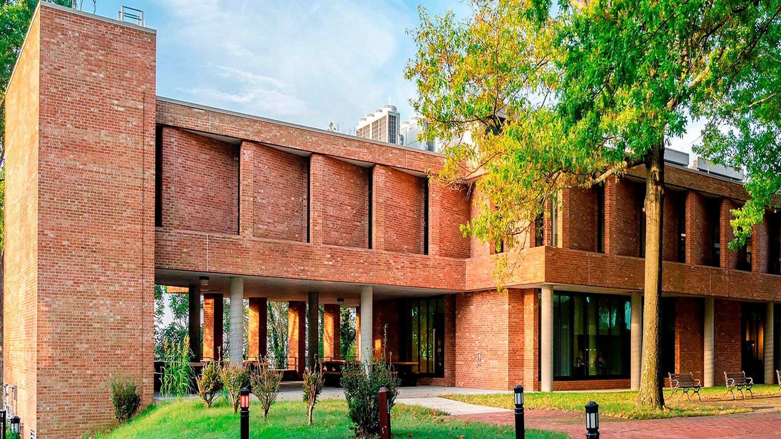 Rectangular brick building. Underneath are round beams with trees and and grass visible beyond.