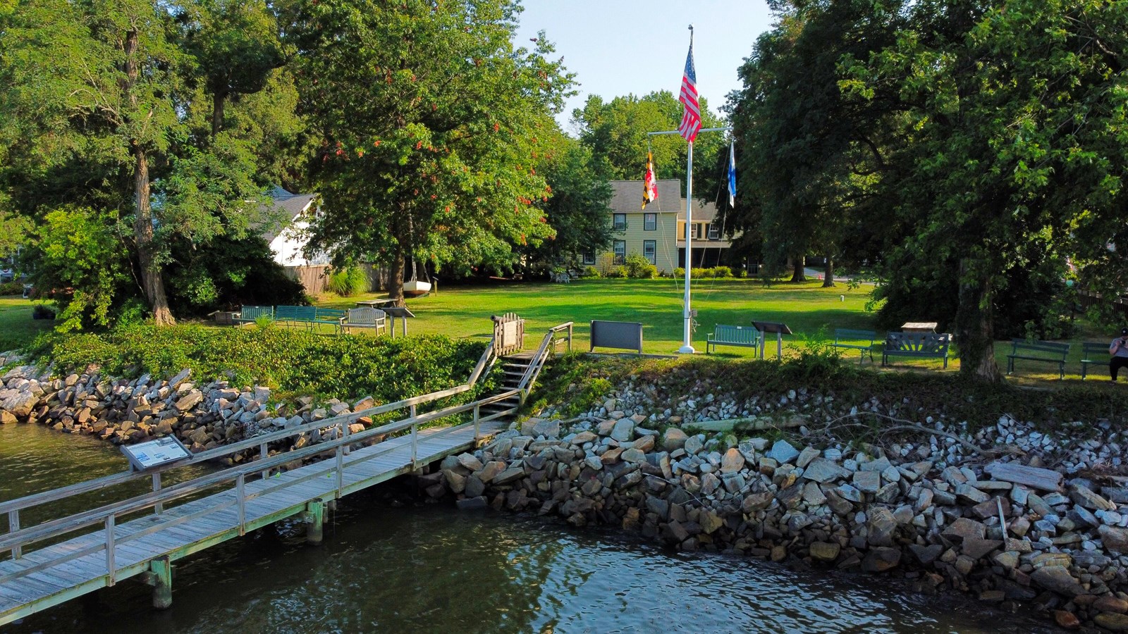 A waterfront scene with a wooden dock, rocky shoreline, grassy area with benches and flagpole.