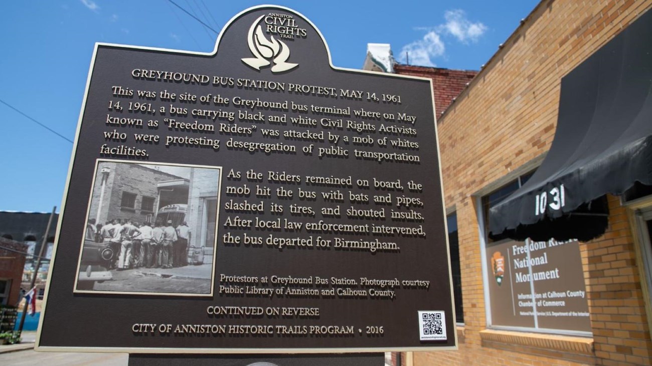 A brown and gold historic marker. In the background: a yellow-brick building with a black awning.