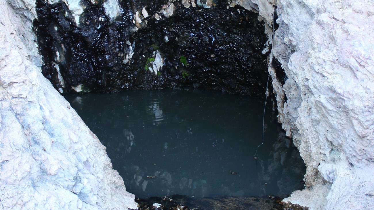 A deep pool of water is nestled in white rocks. The wall behind the water is black from moisture.