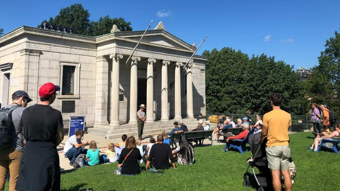 Group of people sitting and standing on grass listening to a Ranger speak outside a granite building