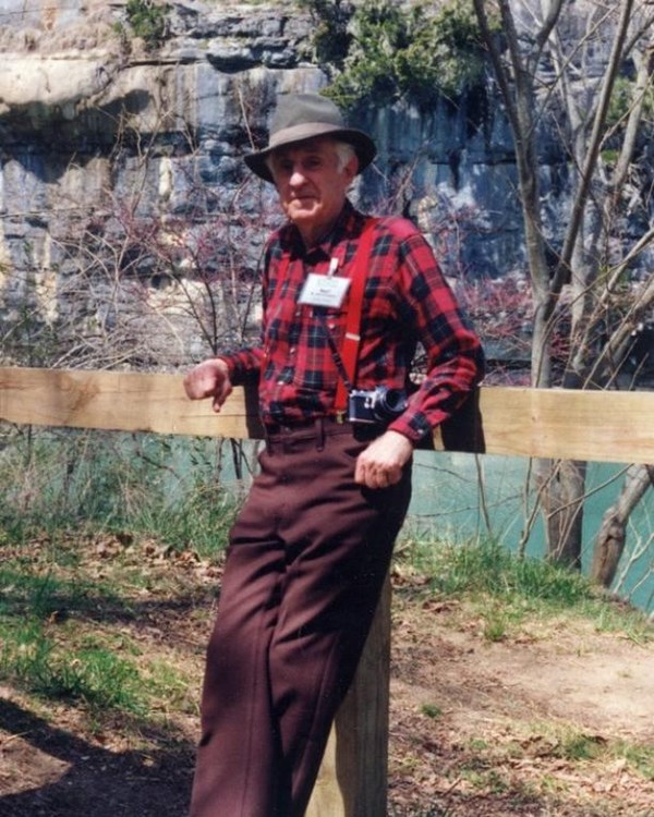 A man wearing a red flannel shirt, maroon pants, and a fedora hat beside the Buffalo River.