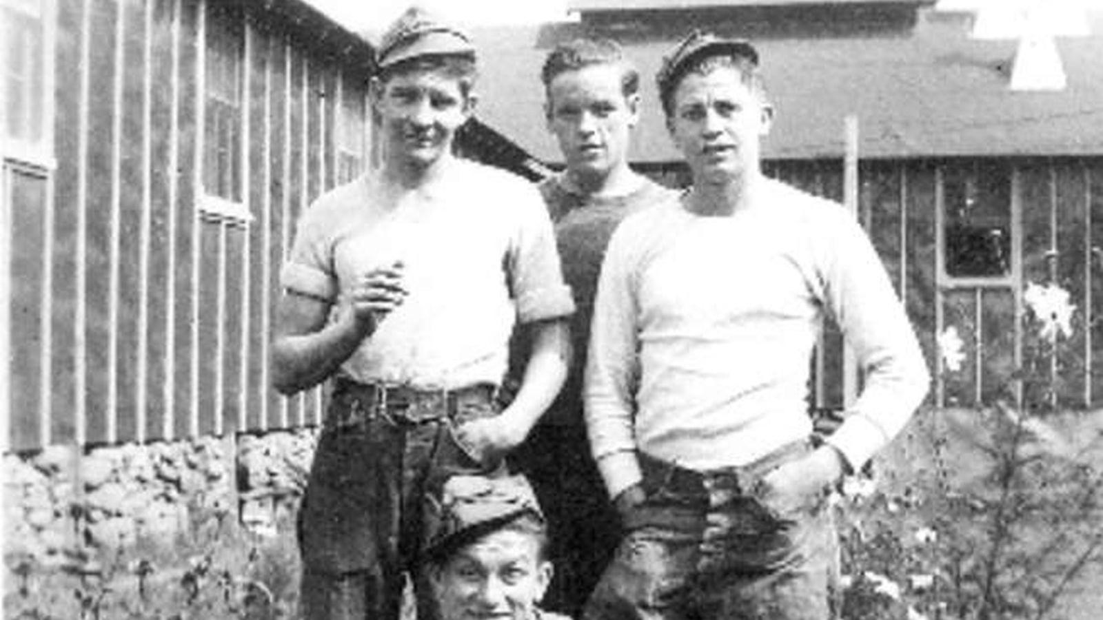 Four CCC boys in their work uniforms pose by the flower bed next to buildings in camp.