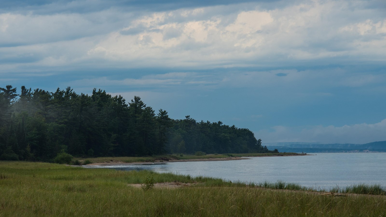 Set back from the lake, a vast patch of grass grows, dark evergreens line the shore in the distance.