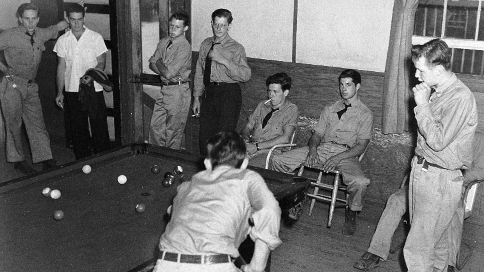  A young man holding a billiard cue is about to make a shot as eight other men look on.