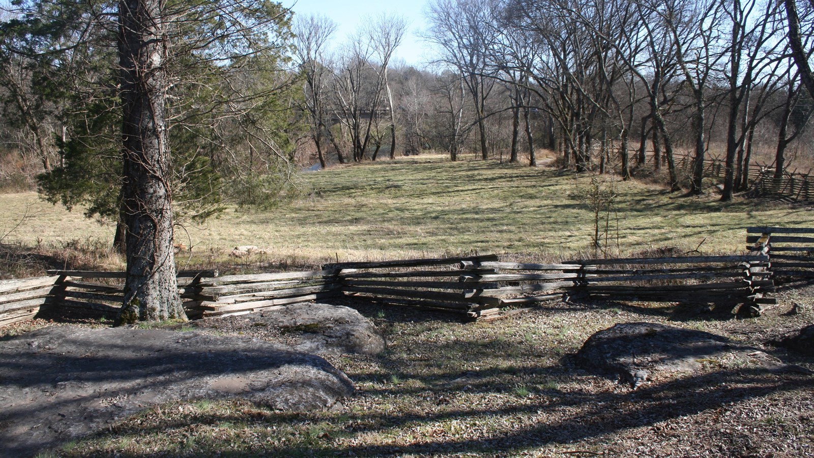 A cedar rail fence cuts across a grass covered slope. A river runs at the base of the slope.