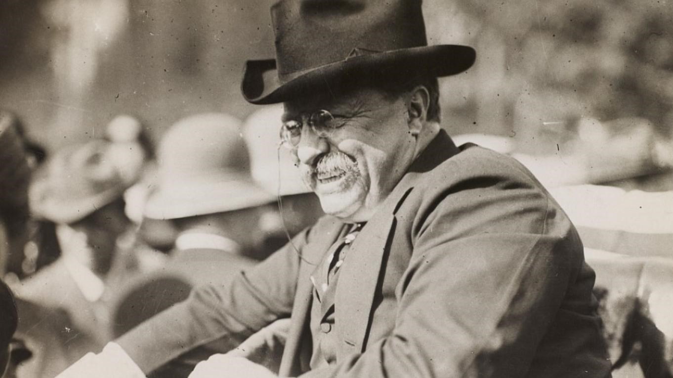 A black-and-white photo of Theodore Roosevelt smiling from an automobile.