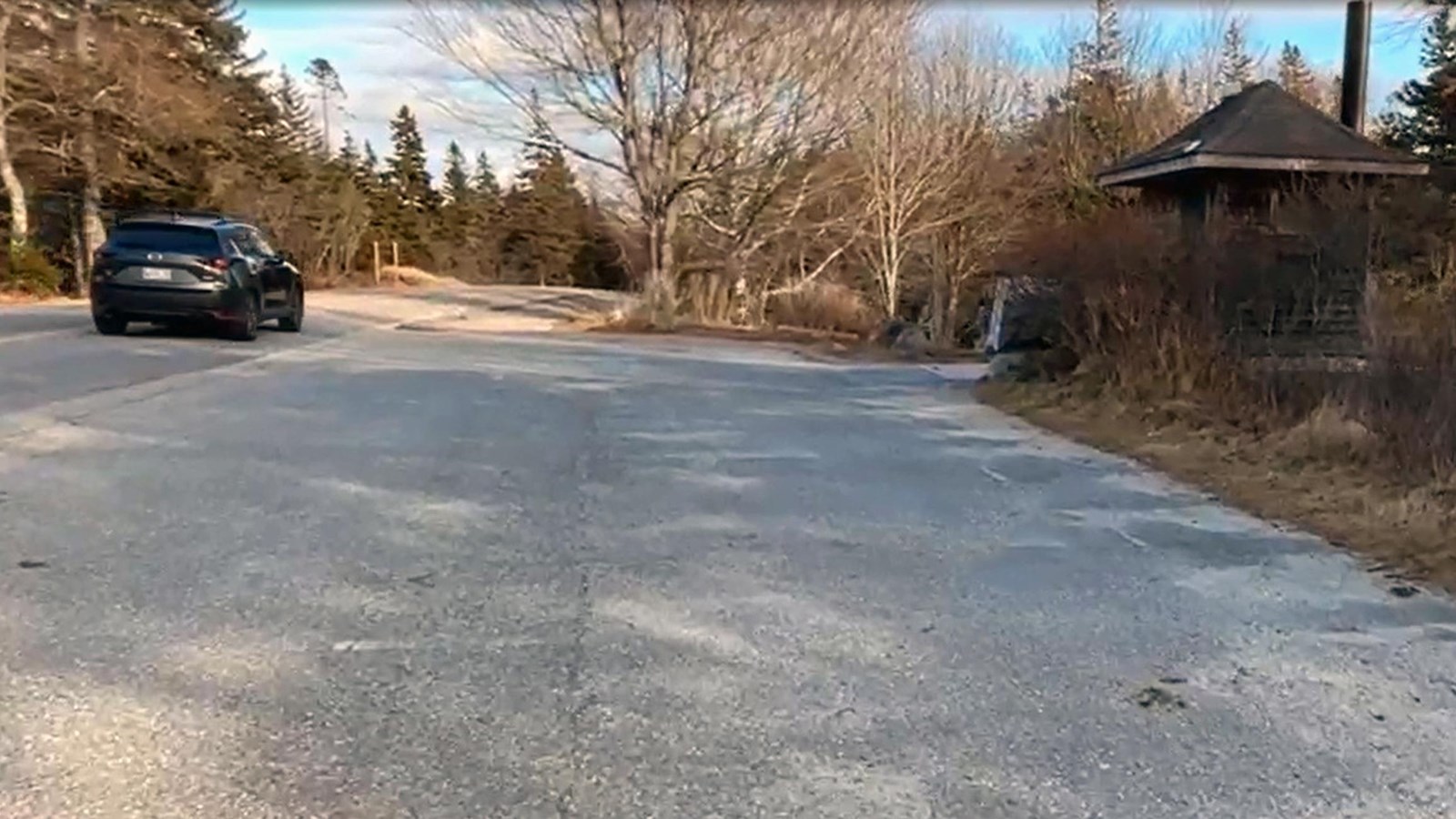 The length of a narrow asphalt parking lot runs along a two-lane road with a black passing car