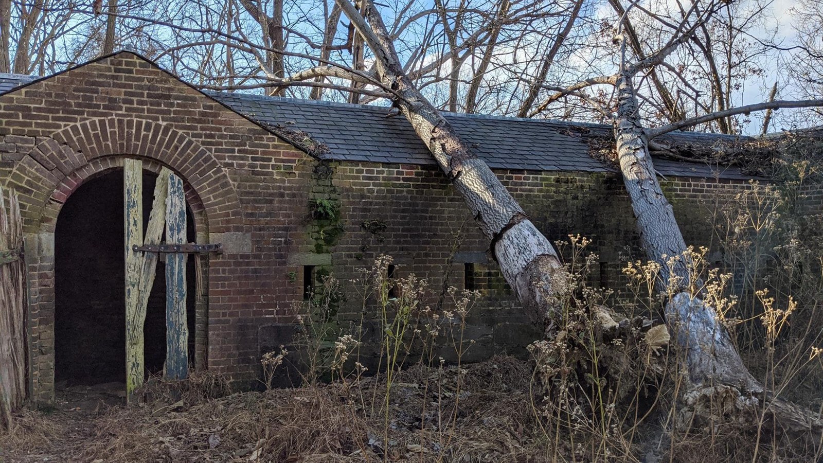 A brick structure with a slate roof. The arched doorway is partially blocked by wooden planks