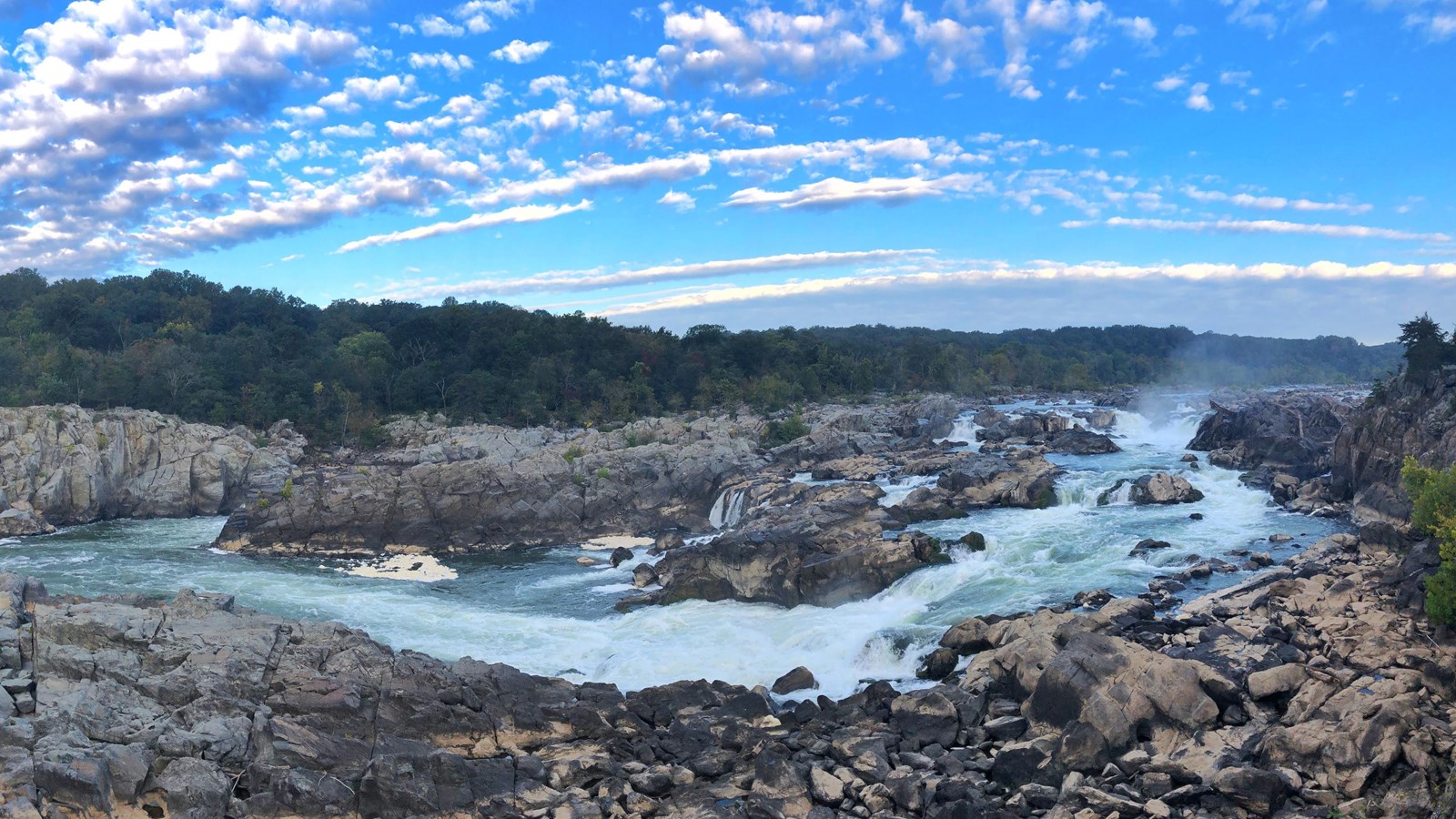 Large, wide waterfall with rocky shorelines lined with trees, on a sunny day