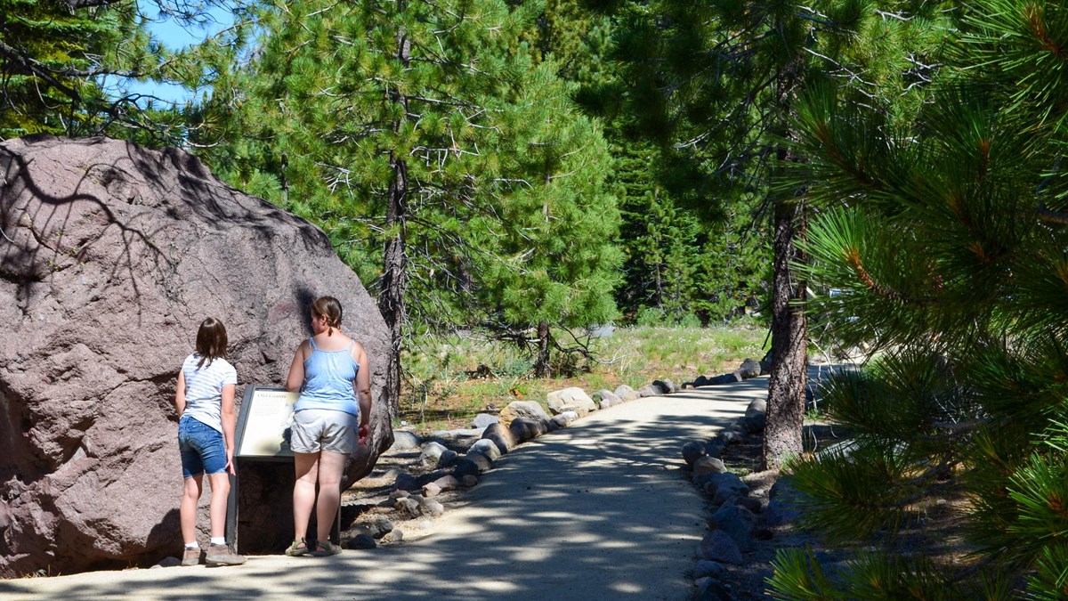 A girl and a woman stand next to a boulder and interpretive sign on a walkway.