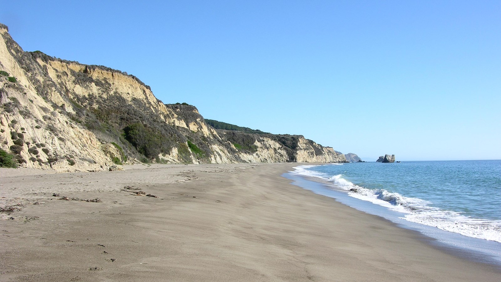 A wave washes in from the right onto a sandy beach at the base of beige-colored bluffs on the left.