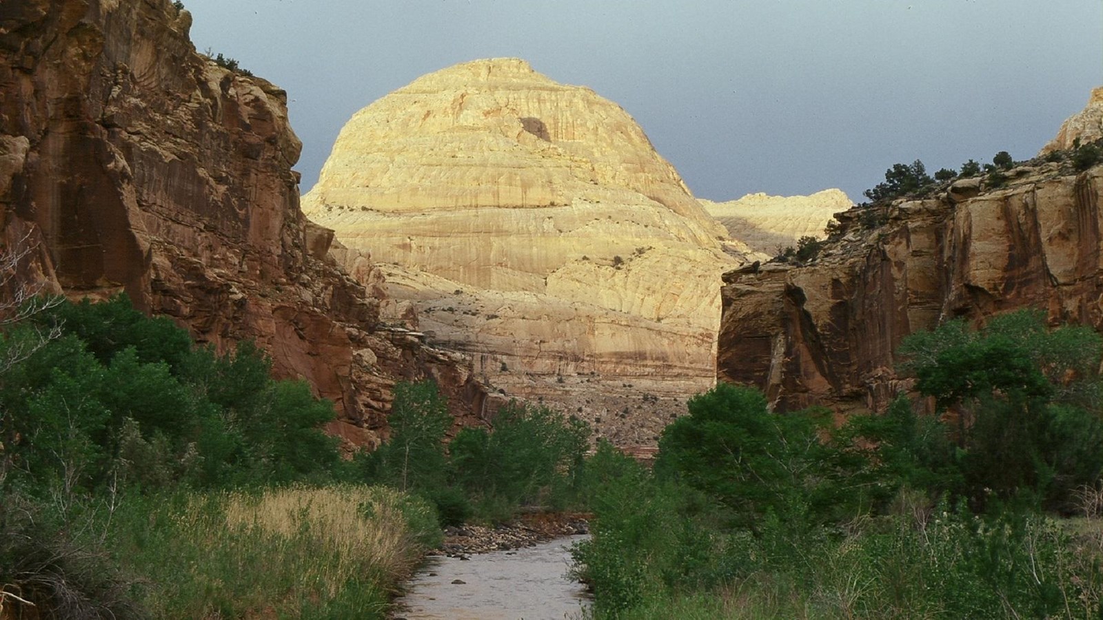 Large rounded white sandstone dome centered above a river, with green grass, red cliffs, & blue sky