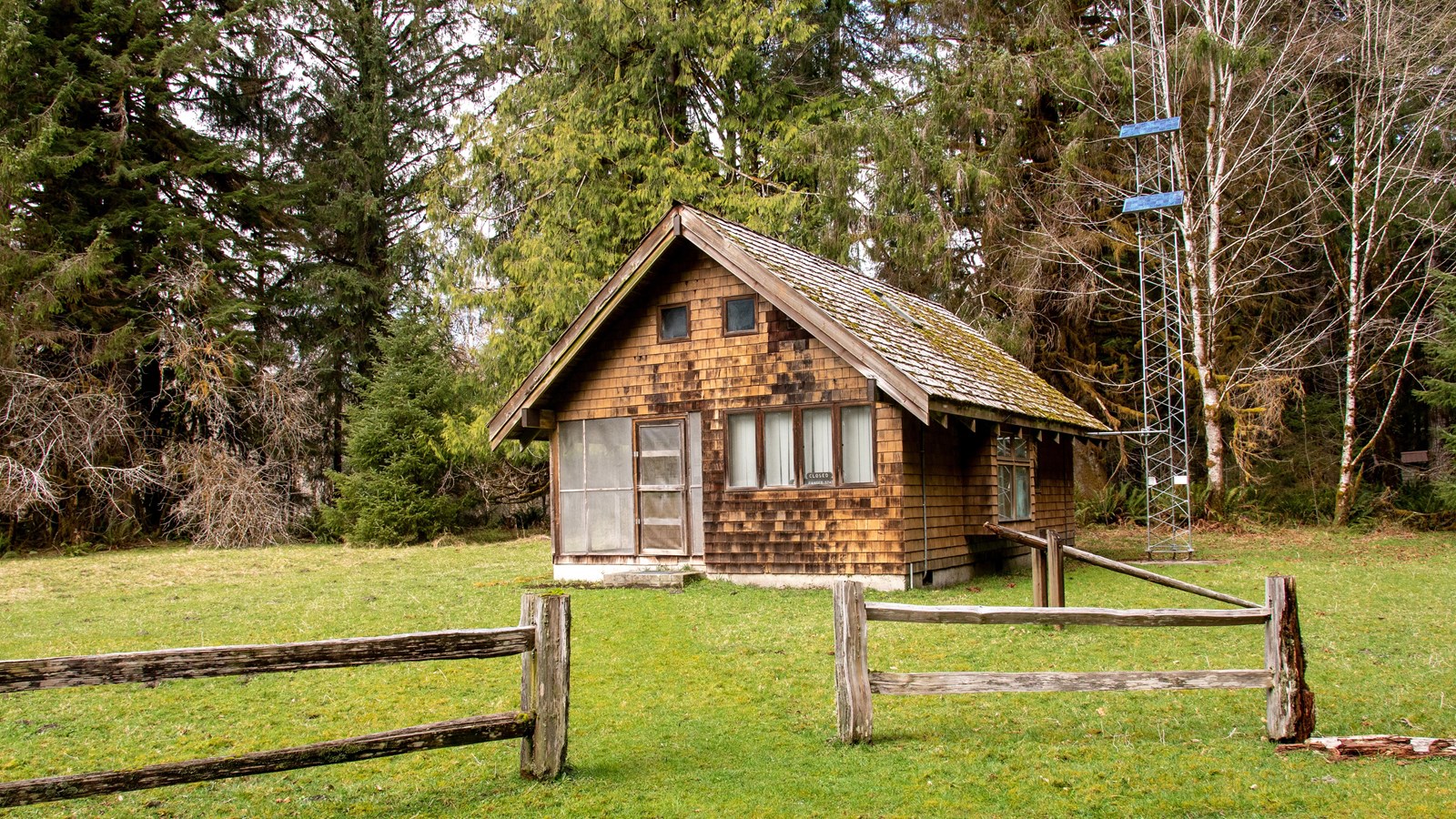 A wooden cabin with a split-rail wooden fence, and tall trees beyond.