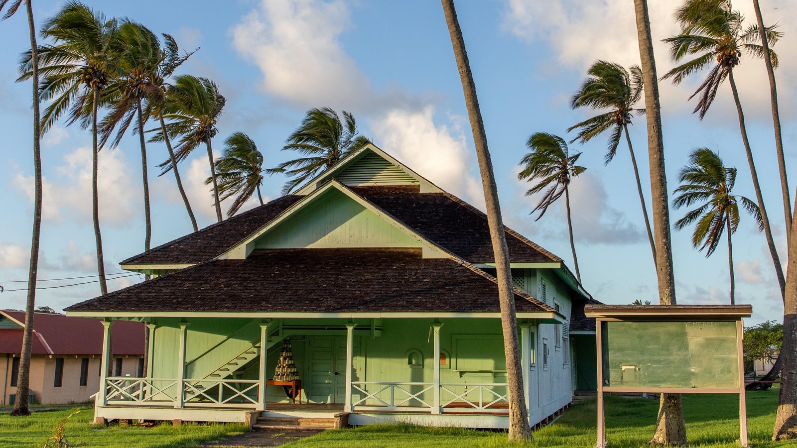 A mint green building with a black roof surrounded by palms trees and a large chalk board.