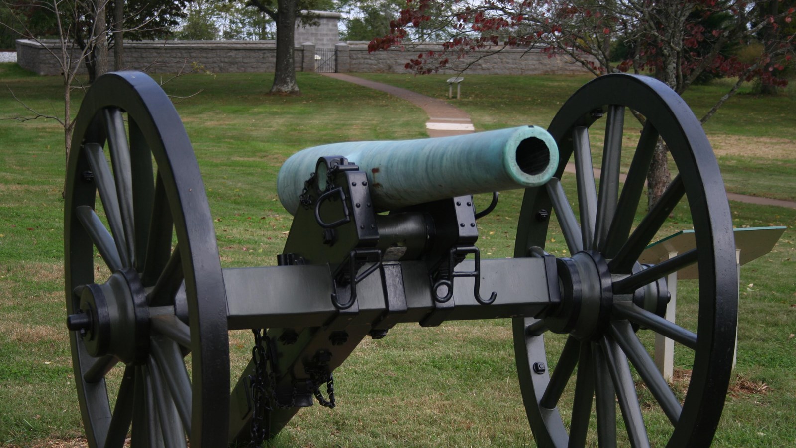 A green colored cannon faces the viewer. a cube shaped monument behind a stone wall in the distance.